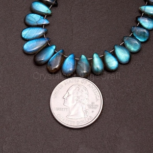May include: A strand of teardrop-shaped labradorite beads in shades of blue and teal. A U.S. quarter coin is placed next to the beads for size comparison. The beads have a polished, iridescent finish. The text "CrystalSageCraft" is visible.
