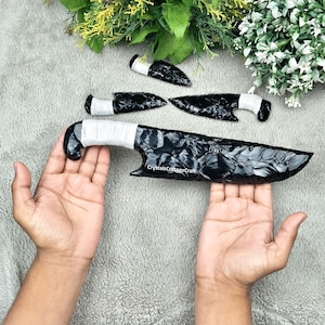 Op de afbeelding: Een verzameling zwarte obsidiaanmessen en pijlpunten met wit omwikkelde handvatten. Het grootste mes wordt in de handen van een persoon gehouden. De obsidiaan heeft een glanzend, reflecterend oppervlak. De achtergrond omvat groen.