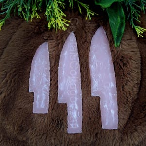 Könnte beinhalten: Drei rosafarbene Pfeilspitzen mit einem weißen Marmormuster. Die Pfeilspitzen sind vertikal auf einer braunen, flauschigen Oberfläche angeordnet.