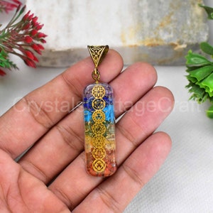 May include: A gold-toned pendant with a rectangular, clear resin inlay featuring seven chakra symbols and corresponding coloured gemstones. The pendant is attached to a gold-toned bail.