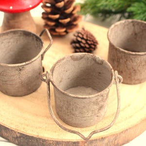 Könnte beinhalten: Drei kleine, rustikale Metallbehälter mit verwittertem Finish. Zwei sind Eimer mit Griffen, einer ist eine Tasse. Die Behälter stehen auf einer Holzscheibe, mit Tannenzapfen und einem Pilz im Hintergrund. Die Behälter haben einen Durchmesser von ca. 7,6 cm.