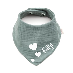 Könnte beinhalten: Ein salbeigrünes Baby-Lätzchen mit dreieckiger Form und Druckknopfverschluss. Das Lätzchen hat drei weiße Herzen und den Namen "Fritzi" in Schreibschrift. Es besteht aus einem weichen, strukturierten Stoff.