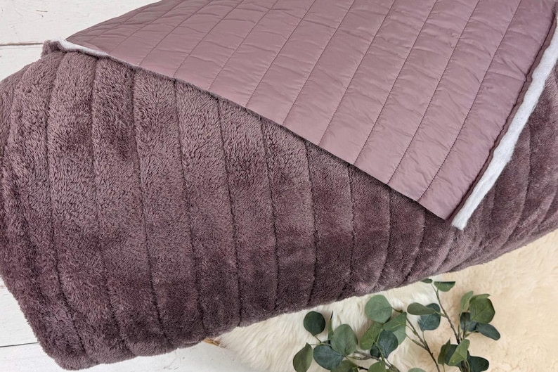 May include: A soft, plush throw blanket in a mauve color with a quilted, mauve-colored backing. The blanket is folded in half and the backing is visible.