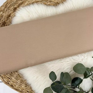 Puede incluir: Primer plano de una muestra de tela marr&oacute;n claro. La tela es lisa y tiene un brillo sutil. Se muestra sobre una alfombra de piel sint&eacute;tica blanca con una cesta tejida debajo.
