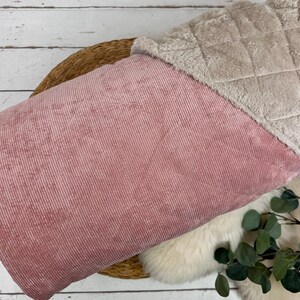 K&ouml;nnte beinhalten: Ein rosa Cordstoff mit einem weichen, gesteppten, beigefarbenen Kunstfellbesatz. Der Stoff liegt auf einem geflochtenen Korb und einer wei&szlig;en, flauschigen Oberfl&auml;che. Gr&uuml;ne Bl&auml;tter sind sichtbar.