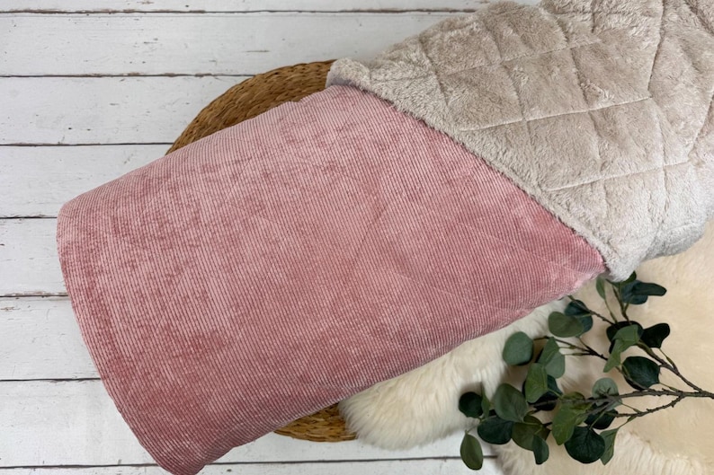K&ouml;nnte beinhalten: Ein rosa Cordstoff mit einem weichen, gesteppten, beigefarbenen Kunstfellbesatz. Der Stoff liegt auf einem geflochtenen Korb und einer wei&szlig;en, flauschigen Oberfl&auml;che. Gr&uuml;ne Bl&auml;tter sind sichtbar.