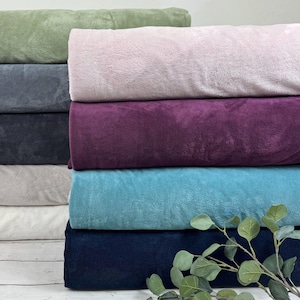 May include: A stack of six fabric swatches in various colors, including green, pink, purple, blue, and white. The swatches are folded and arranged in a pile, with a sprig of green leaves in front.