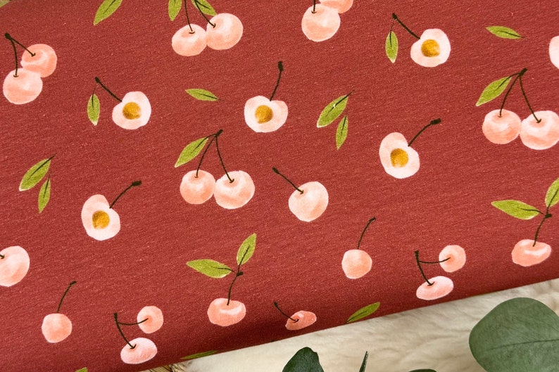May include: A red fabric with a repeating pattern of pink cherries with green stems and leaves.