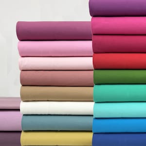 Puede incluir: Una pila de muestras de tela en varios colores, incluyendo morado, rosa, rojo, verde, azul y beige. La tela está doblada cuidadosamente y dispuesta en una pila vertical.