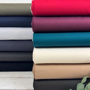 Peut inclure: Une pile de rouleaux de tissu de différentes couleurs, dont vert olive, bleu marine, gris clair, sarcelle, crème et marron. Les rouleaux sont soigneusement disposés, mettant en valeur les différentes nuances et textures du matériau. Une brindille de verdure est au premier plan.