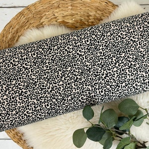 May include: Cream-coloured fabric with a black leopard print pattern. The fabric is rolled up and displayed on a woven basket.