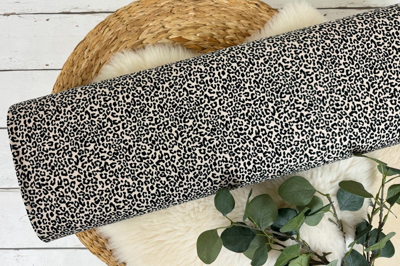 May include: Cream-coloured fabric with a black leopard print pattern. The fabric is rolled up and displayed on a woven basket.