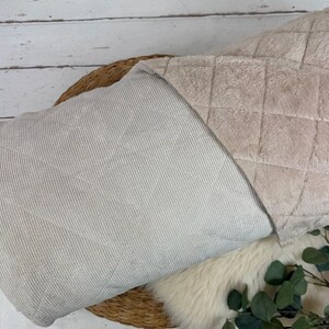 K&ouml;nnte beinhalten: Eine beige Decke mit einer gesteppten Oberseite und einer gestreiften Unterseite. Die Decke ist &uuml;ber einem Weidenkorb gefaltet, mit einer flauschigen wei&szlig;en Oberfl&auml;che und Gr&uuml;n im Vordergrund. Die Abmessungen der Decke sind nicht sichtbar.
