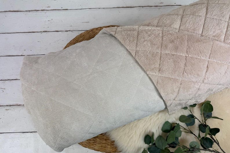 K&ouml;nnte beinhalten: Eine beige Decke mit einer gesteppten Oberseite und einer gestreiften Unterseite. Die Decke ist &uuml;ber einem Weidenkorb gefaltet, mit einer flauschigen wei&szlig;en Oberfl&auml;che und Gr&uuml;n im Vordergrund. Die Abmessungen der Decke sind nicht sichtbar.