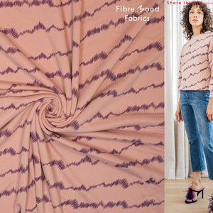 Op de afbeelding: Roze stof met een patroon van paarse golvende lijnen. De stof is gevouwen en gedraaid. De tekst "Fibre Good Fabrics" is zichtbaar in de rechterbovenhoek van de afbeelding.