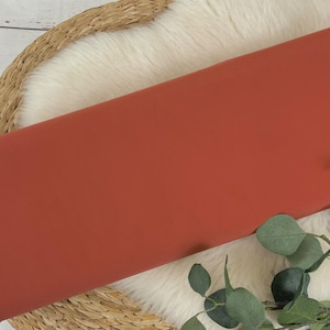 May include: A close-up of a rust-colored fabric swatch. The fabric is smooth and has a matte finish. It is lying on a white faux fur rug with a woven basket underneath.