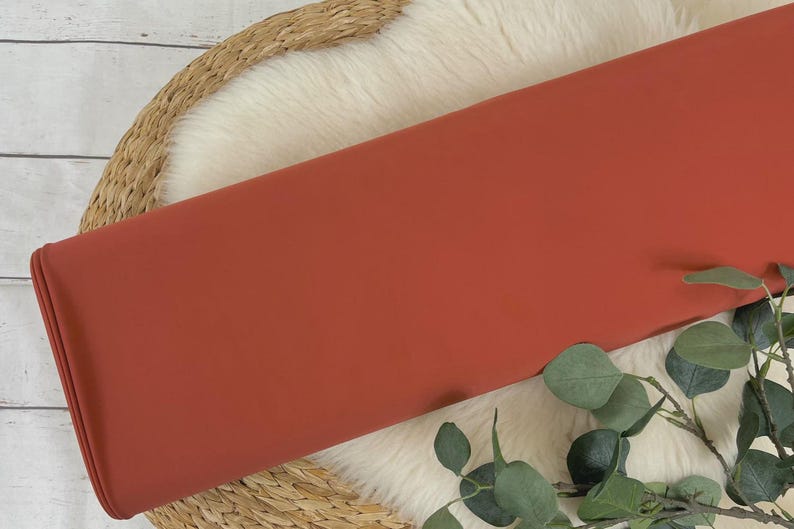 May include: A close-up of a rust-colored fabric swatch. The fabric is smooth and has a matte finish. It is lying on a white faux fur rug with a woven basket underneath.