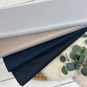 Può includere: Pile di rotoli di tessuto in vari colori: grigio chiaro, beige, blu navy e nero. I tessuti sono disposti ordinatamente su una superficie di legno bianco, con un rametto di foglie verdi e un tappeto di pelliccia bianca sullo sfondo.