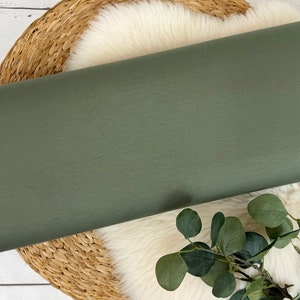 May include: A roll of olive green fabric rests on a woven basket and a white, fluffy surface. The fabric is a solid color and appears to be a medium-weight material. Greenery accents the scene.