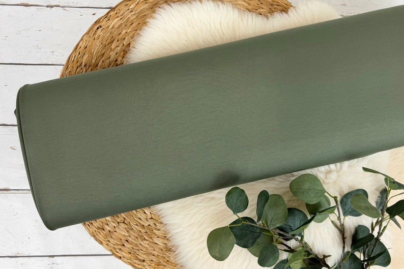 May include: A roll of olive green fabric rests on a woven basket and a white, fluffy surface. The fabric is a solid color and appears to be a medium-weight material. Greenery accents the scene.