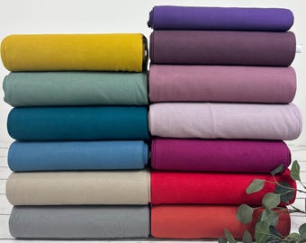 Könnte beinhalten: Ein Stapel Stoffrollen in verschiedenen Farben, darunter Senfgelb, Türkis und Altrosa. Die Rollen sind ordentlich angeordnet und zeigen die verschiedenen Farbtöne und Texturen des Materials. Das Bild zeigt die Vielfalt der verfügbaren Farben.
