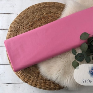 May include: A pink fabric swatch displayed on a woven mat with a white faux fur rug and green foliage.