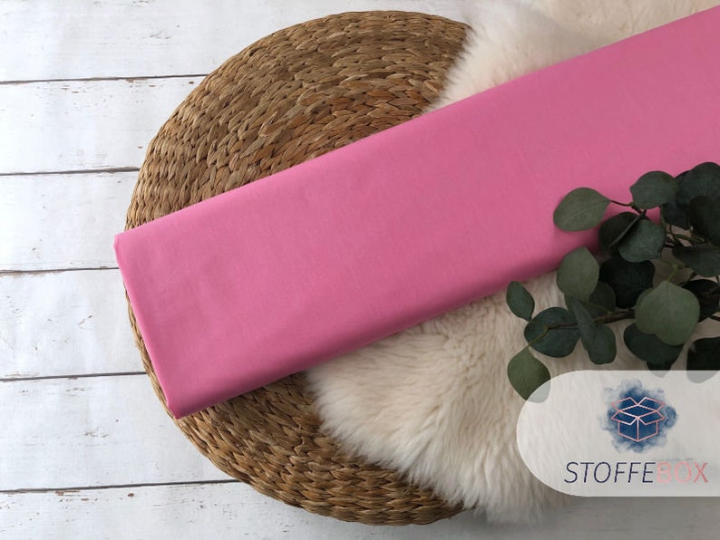 May include: A pink fabric swatch displayed on a woven mat with a white faux fur rug and green foliage.