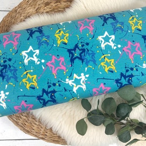 May include: Blue fabric with a white, yellow, pink, and blue star pattern. The stars are outlined in black and have a paint splatter effect.