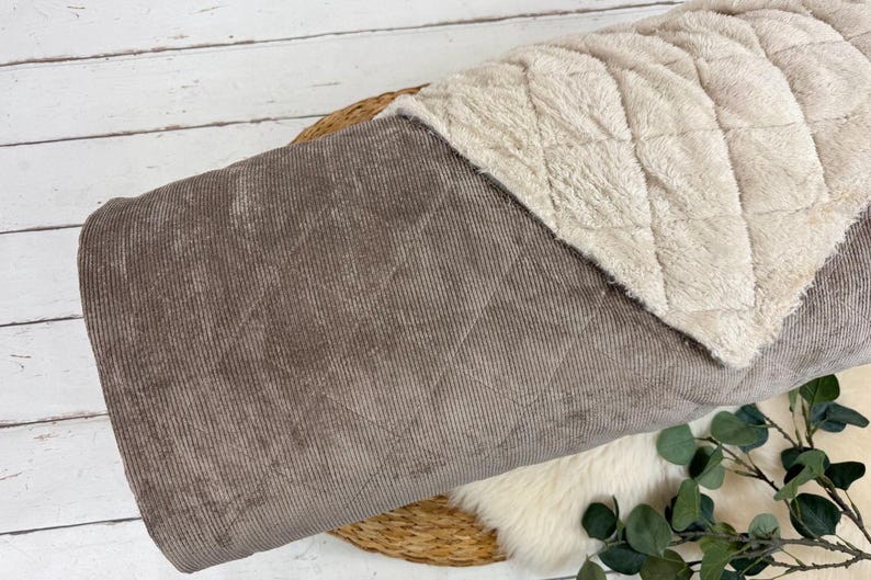 K&ouml;nnte beinhalten: Eine gerollte Decke mit zweifarbigem Design. Die Au&szlig;enseite besteht aus braunem Cord, w&auml;hrend die Innenseite aus weichem, cremefarbenem, gestepptem Stoff besteht. Die Decke liegt auf einer gewebten Matte und einer wei&szlig;en Holzoberfl&auml;che.