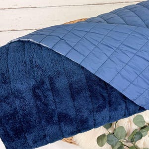 May include: A blue quilted blanket with a soft, plush side. The blanket is folded in half and is lying on a white wooden surface.