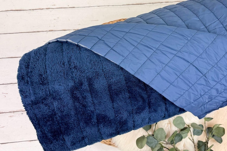 May include: A blue quilted blanket with a soft, plush side. The blanket is folded in half and is lying on a white wooden surface.