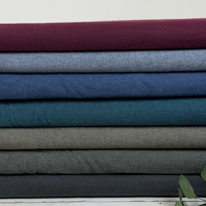 Op de afbeelding: Een stapel stof in verschillende kleuren, waaronder bordeauxrood, spijkerblauw, blauwgroen en houtskool. De stof lijkt een zacht, getextureerd materiaal te zijn, mogelijk flanel of fleece. De vouwen van de stof zijn zichtbaar, waardoor een gelaagd effect ontstaat.