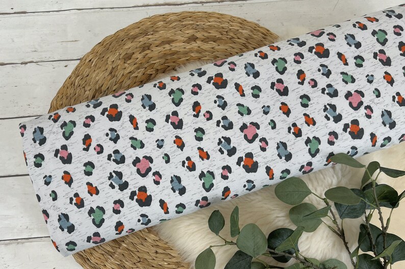 May include: White fabric with a colourful leopard print pattern. The print features black spots with accents of orange, pink, and green.