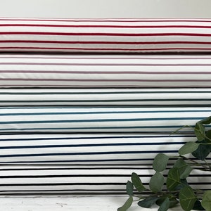 Op de afbeelding: Een stapel van vijf gestreepte stofrollen in verschillende kleuren, waaronder rood, roze, groen, blauw en zwart. De stof is wit met gekleurde strepen.