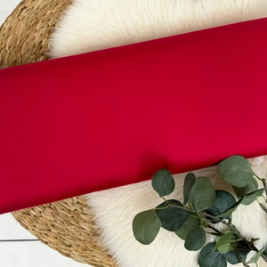 May include: A roll of vibrant red fabric, likely textile material, is displayed. The fabric is smooth and appears to be a solid color. The roll rests on a white, fluffy surface, with a woven basket and green foliage in the background.