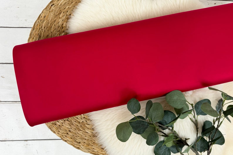 May include: A roll of vibrant red fabric, likely textile material, is displayed. The fabric is smooth and appears to be a solid color. The roll rests on a white, fluffy surface, with a woven basket and green foliage in the background.