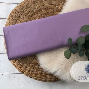 May include: A piece of lavender-colored fabric is displayed on a woven straw mat. The fabric is smooth and has a subtle sheen. A sprig of green foliage is placed on the fabric.