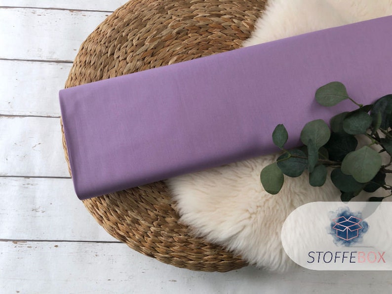 May include: A piece of lavender-colored fabric is displayed on a woven straw mat. The fabric is smooth and has a subtle sheen. A sprig of green foliage is placed on the fabric.