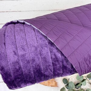 May include: A purple reversible blanket with a soft, plush side and a quilted side. The blanket is rolled up and partially unfurled, revealing both sides.