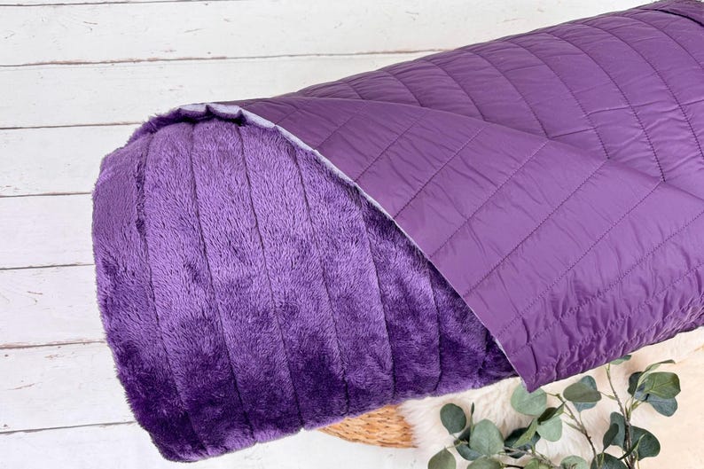May include: A purple reversible blanket with a soft, plush side and a quilted side. The blanket is rolled up and partially unfurled, revealing both sides.