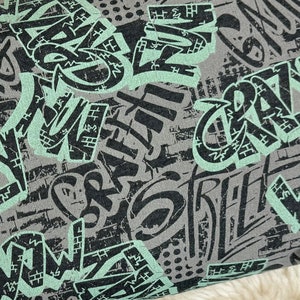 Könnte beinhalten: Grauer und grüner Stoff mit einem sich wiederholenden Muster aus Graffiti-artigen Wörtern wie "Crazy", "Street" und "Wow".