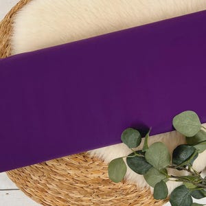 May include: A solid purple fabric swatch displayed on a woven basket with a white faux fur lining and eucalyptus leaves.