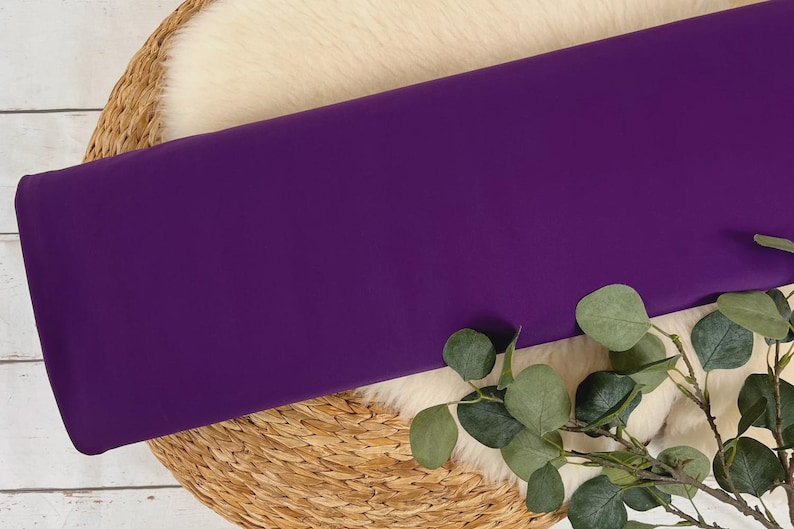 May include: A solid purple fabric swatch displayed on a woven basket with a white faux fur lining and eucalyptus leaves.