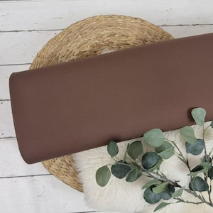 May include: A roll of solid brown fabric rests on a woven basket and a white fluffy surface, with a sprig of green leaves in the foreground. The fabric is a rich, warm brown color, and the overall aesthetic is natural and earthy.