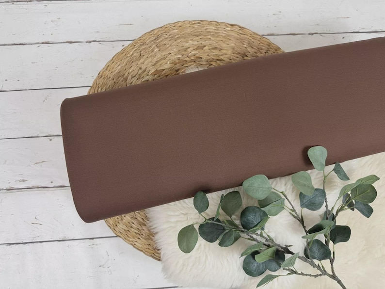 May include: A roll of solid brown fabric rests on a woven basket and a white fluffy surface, with a sprig of green leaves in the foreground. The fabric is a rich, warm brown color, and the overall aesthetic is natural and earthy.