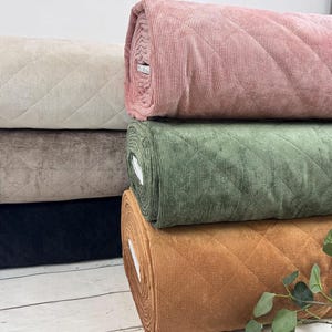 K&ouml;nnte beinhalten: Eine Auswahl an Stoffrollen in verschiedenen Farben, darunter Beige, Braun, Schwarz, Rosa, Gr&uuml;n und Orange. Der Stoff hat ein gestepptes Rautenmuster und eine Cordstruktur. Ein Zweig mit gr&uuml;nen Bl&auml;ttern ist im Vordergrund zu sehen.