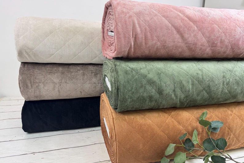 K&ouml;nnte beinhalten: Eine Auswahl an Stoffrollen in verschiedenen Farben, darunter Beige, Braun, Schwarz, Rosa, Gr&uuml;n und Orange. Der Stoff hat ein gestepptes Rautenmuster und eine Cordstruktur. Ein Zweig mit gr&uuml;nen Bl&auml;ttern ist im Vordergrund zu sehen.