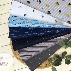 Op de afbeelding: Een stapel van zes verschillende stoffen met maritieme thema's. De prints omvatten een blauw-wit gestreept patroon, een blauw-wit ankerpatroon, een blauw-wit golfpatroon, een blauw-wit vispatroon, een wit-blauw zeilbootpatroon en een grijs-wit piratenpatroon. De stof wordt getoond op een geweven mand.