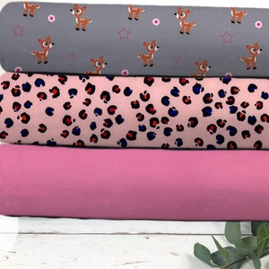 Könnte beinhalten: Drei Stoffrollen, eine mit rosa Hintergrund und Leopardenmuster, eine mit grauem Hintergrund und weißem Hirschmuster und eine einfarbig rosa.