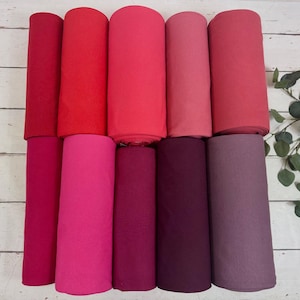 Pode incluir: Uma coleção de rolos de tecido em vários tons de vermelho e rosa. Os rolos são dispostos verticalmente, mostrando um gradiente de vermelho escuro a rosa claro. O tecido parece ser de cor sólida, com uma textura suave. Um raminho de vegetação está no fundo.